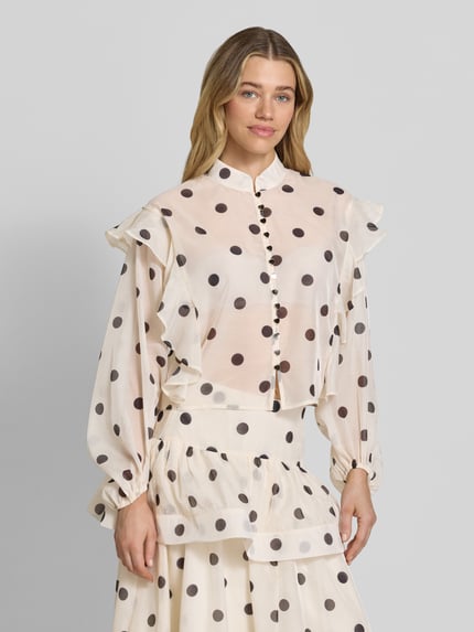 Frauenmodel trägt Boxy Fit Chiffon-Bluse mit Polka-Dots-Muster Modell 'DOROTHY' von Vero Moda, semitransparent mit Rüschen-Applikation, Allover-Muster, Knopfleiste, Ballonärmel, Stoffband, helle Grundfarbe, fließende Textur, Ganzkörperansicht von vorne.
