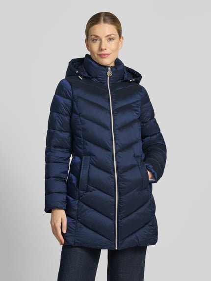 Frauenmodel trägt Steppmantel mit Kapuze und Reißverschlusstaschen von MICHAEL Michael Kors in Dunkelblau, glänzende Oberfläche, Ganzkörperansicht von vorne. Gerader Schnitt, Kapuze mit Tunnelzug, seitliche Reißverschlusstaschen, sichtbare Steppungen, Kontext: Rückenlänge 86 cm, Schulterbreite 43 cm, Brustweite 50 cm, Ärmellänge 62 cm.