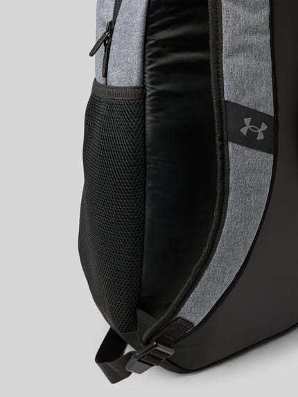 Produktfoto, Rugzakfunctie met draagband en verstelbare schouderband van Under Armour, hoofvak met ritssluiting, labelprint, zijvak met ritssluiting, grijs en zwart, breedte 31 cm, lengte 48 cm, diepte 18 cm, bandlengte 53 cm.