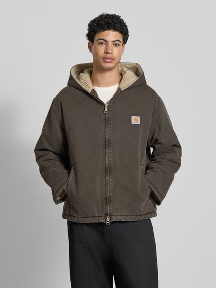 Männermodel trägt Jacke mit Zweiwege-Reißverschluss Modell 'MITCH' von Carhartt aus brauner Baumwolle mit Kapuze und Label-Detail, Ganzkörperansicht von vorne.