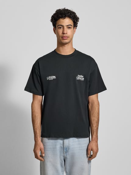 Männermodel trägt T-Shirt mit Label-Print von ANOTHER COTTON LAB aus reiner Baumwolle, lockerer Schnitt, gerippter Rundhalsausschnitt, Ganzkörperansicht von vorne.