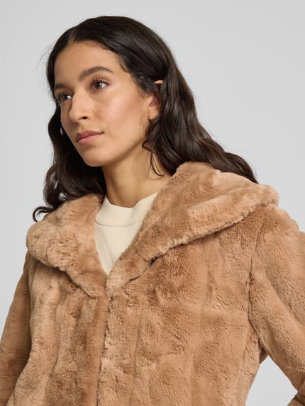 Frauenmodel trägt Jacke aus Kunstfell Modell 'NEW SOPHY' von Guess in Beige mit Schalkragen, Regular Fit, Nahaufnahme Oberkörper. Rückenlänge 56 cm, Schulterbreite 44 cm, Brustweite 46 cm, Ärmellänge 59 cm.