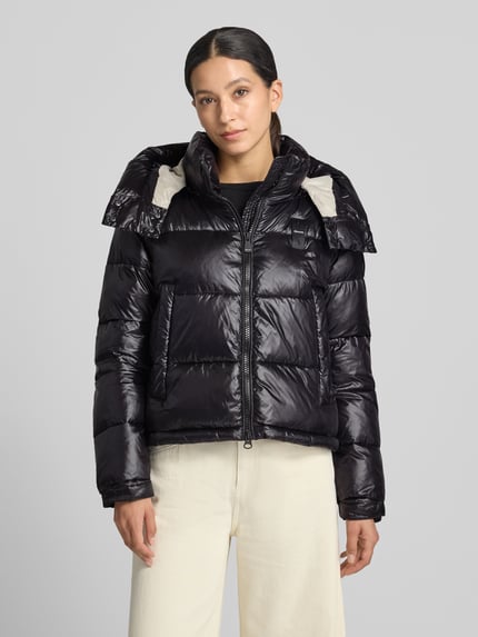Frauenmodel trägt Regular Fit Jacke mit Sorona-Füllung Modell 'GLORIA' von Blauer USA in Schwarz, glänzende Stepp-Optik, Stehkragen mit Kapuze, Eingrifftaschen, Zweiwege-Reißverschluss, Logo-Detail, Ganzkörperansicht von vorne.