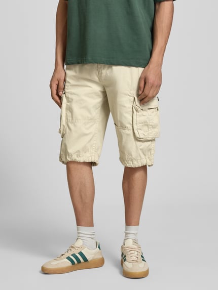 Männermodel trägt Bermudas mit Pattentaschen und Logo-Stitching von Alpha Industries in Beige, Reine Baumwolle, Straight Fit, Ganzkörperansicht von vorne.