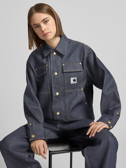 Frauenmodel trägt Regular Fit Jeansjacke mit Logo-Patch Modell 'Arctic' von Carhartt Work In Progress aus reiner Baumwolle in Dunkelblau, goldene Knöpfe, Umlegekragen, Brust- und Eingrifftaschen, Nahaufnahme Oberkörper.