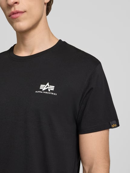 Männermodel trägt T-Shirt mit Logo und Rundhalsausschnitt von Alpha Industries aus Reine Baumwolle im Regular-Fit, Logo vorn, Nahaufnahme Oberkörper. Rückenlänge bei Größe M: 67 cm.