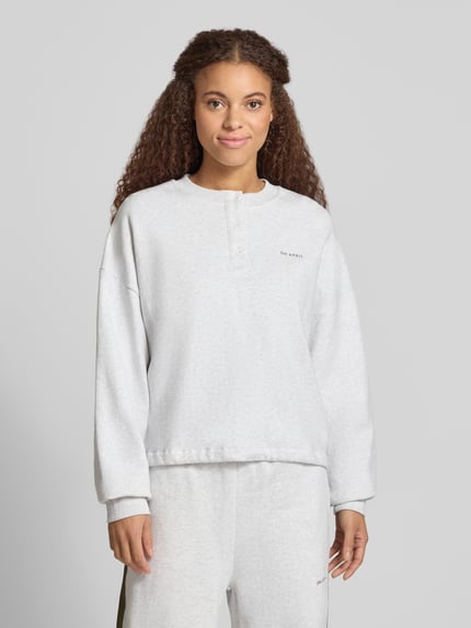 Kobieta modelka nosi Boxy Fit Sweatshirt mit Knopfleiste Modell 'Aven' od OH APRIL z bawełny, okrągły dekolt, wyhaftowane logo, pudełkowy krój, jasnoszara tkanina o gładkiej fakturze, widok całej sylwetki z przodu.