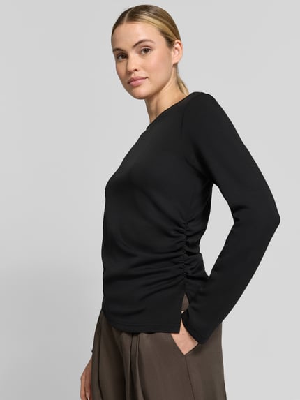Modelka nosi czarną bluzkę z długim rękawem Regular Fit Longsleeve mit Raffungen Modell 'Helle' von My Essential Wardrobe, elastyczna mieszanka materiałów z udziałem modalu, marszczenia, dekolt w łódkę, długość tyłu w rozmiarze S 62 cm, widok całej sylwetki z przodu.