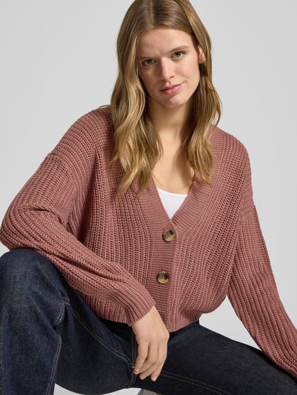 Kobiece model nosi Regular Fit Strickjacke mit einreihiger Knopfleiste Modell 'CAROL NICE' od Only, róż jednolity, krój regular fit, głęboki dekolt w serek, długi rękaw, dzianinowa faktura, widoczny ścieg, wersja cropped, przód, zbliżenie na górną część ciała.