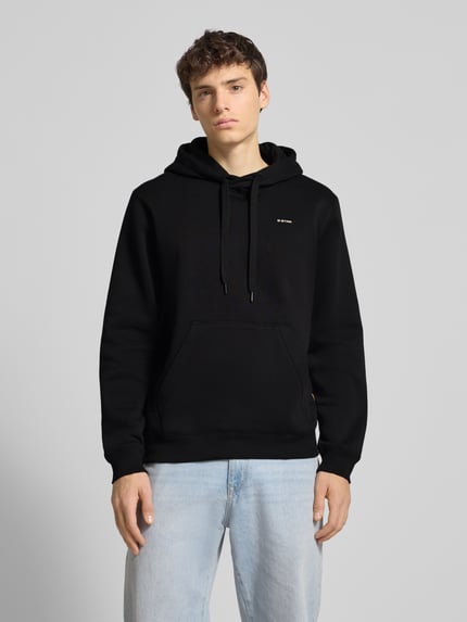 Männermodel trägt Hoodie mit Kapuze von G-Star Raw aus schwarzem Baumwoll-Mix, Label-Print, Kängurutasche, lockerer Schnitt, Ganzkörperansicht von vorne.