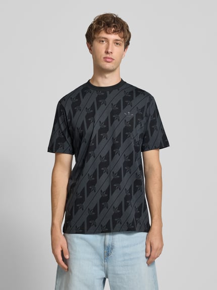 Männermodel trägt T-Shirt aus reiner Baumwolle Modell 'MONOGRAMM AOP' von adidas Originals in Schwarz mit Muster, Ganzkörperansicht von vorne.
