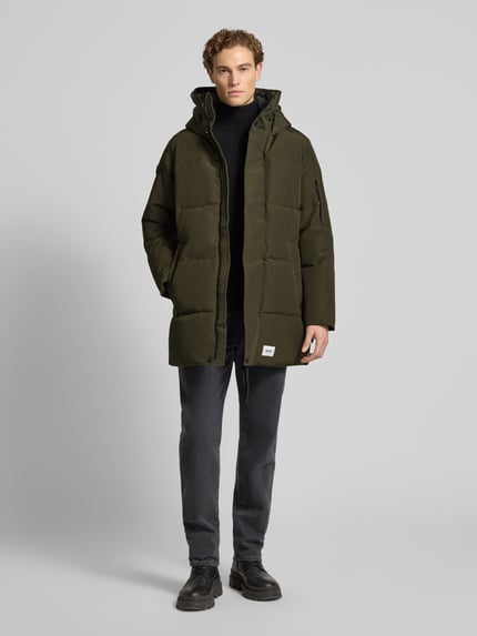 Männermodel trägt Regular Fit Steppmantel mit Logo-Patch Modell 'Zeno' von khujo in Oliv mit Kapuze und elastischen Ärmelabschlüssen, Ganzkörperansicht von vorne.
