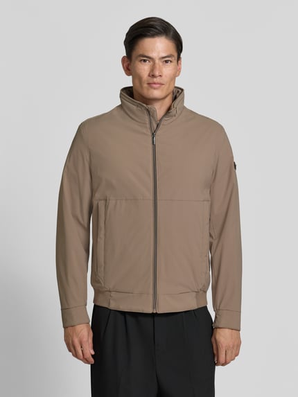Männermodel trägt Regular Fit Jacke mit stójka von bugatti w beżowym kolorze, wpuszczane kieszenie, zamek błyskawiczny, elastyczny pas, długość tyłu w rozmiarze 50: 67 cm, szerokość ramion w rozmiarze 50: 45 cm, szerokość klatki piersiowej w rozmiarze 50: 58 cm, długość rękawów w rozmiarze 50: 67 cm, całkowity widok z przodu