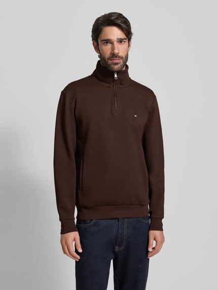 Männermodel trägt Regular Fit Troyer aus Baumwoll-Mix von Tommy Hilfiger, unifarben braun mit Label-Detail, Reißverschluss, Ganzkörperansicht von vorne.