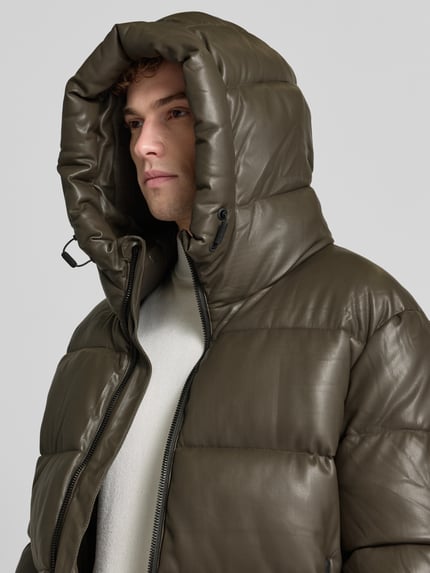 Męski model nosi kurtkę pikowaną z kapturem Model 'COEN 2' od khujo, krój regular fit, detal z zamkiem błyskawicznym, detail z logo, zieleń oliwkowa, perspektywa zbliżenie na górną część ciała. Długość tyłu w rozmiarze M: 72 cm, szerokość ramion w rozmiarze M: 55 cm, szerokość w klatce piersiowej w rozmiarze M: 61 cm, długość rękawa w rozmiarze M: 62 cm