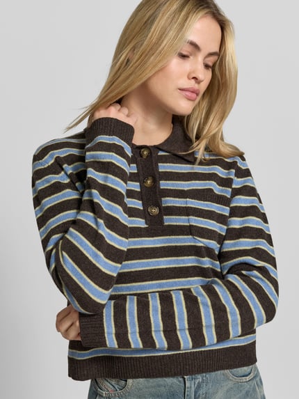Vrouwenmodel draagt Regular Fit poloshirt uit zuivere wol van BAUM & PFERDGARTEN met tricotlook, polokraag, met lange mouwen, gestreept in bruin, blauw en geel, ruglengte bij maat S 51 cm, half close-up bovenlichaam.