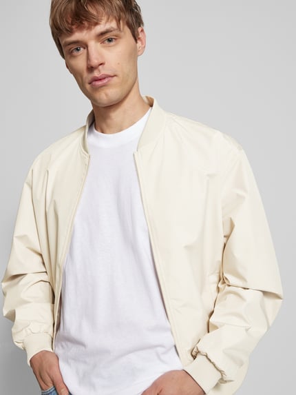 Męski model nosi Bomberjacke mit seitlichen Eingrifftaschen Modell 'CHARGE' od Jack & Jones, krój straight fit, stójka, zamek błyskawiczny, boczne kieszenie, ściągacze, długość tyłu w rozmiarze M 66 cm, szerokość ramion w rozmiarze M 54 cm, szerokość klatki piersiowej w rozmiarze M 65 cm, długość rękawa w rozmiarze M 64 cm, kremowy kolor, prosty fason, matowa faktura, całkowity widok z przodu.