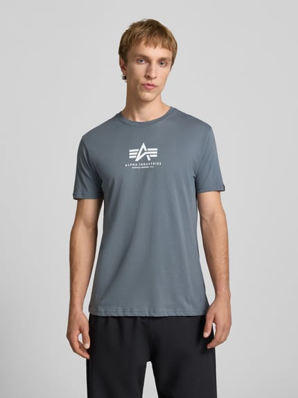 Männermodel trägt T-Shirt mit Logo und Rundhalsausschnitt von Alpha Industries in Grau, reine Baumwolle, Regular Fit, Ganzkörperansicht von vorne.