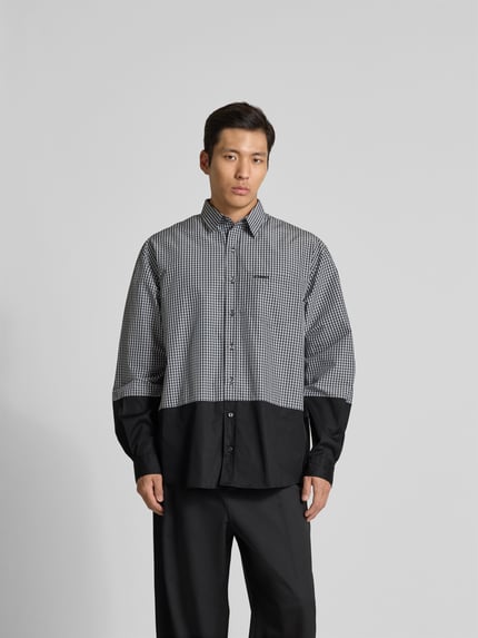 Mężczyznamodel nosi Relaxed Fit Freizeithemd mit Brusttasche von VETEMENTS, mieszanka bawełny, krój relaxed fit, listwa guzikowa na całej długości, brustasza z wyhaftowanym logo, krój z długim rękawem, długość tyłu w rozmiarze M: 85 cm, szerokość w rozmiarze M: 53 cm, długość rękawów w rozmiarze M: 68 cm, czarno-biała kratka z kontrastem, tekstura gładka, widok całej sylwetki od przodu.