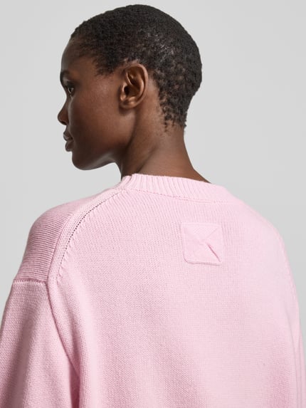 Männermodel trägt Oversized Wollpullover aus Woll-Mix von Nanushka, strickoptik, różowy kolor, karo faktura, stehkrajen, raglanowe rękawy, gerippte ściągacze, mieszanka wełny i bawełny, widok z tyłu, detal na karku, długość tyłu w rozmiarze S: 61 cm. Ganzkörperansicht Rückansicht