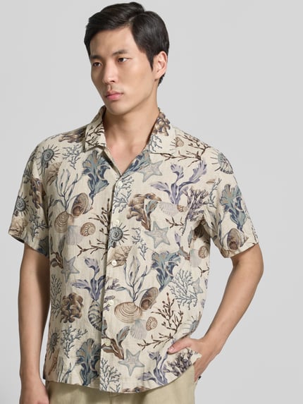 Männermodel trägt Regular Fit Freizeithemd aus reinen Leinen von Gant mit Allover-Motiv-Print, Reverskragen, Logo-Stitching, Kurzärmelig, Knopfleiste und Brusttasche in Naturtönen mit blauen und braunen Akzenten, Textur fein und grob gemustert, Ganzkörperansicht von vorne.