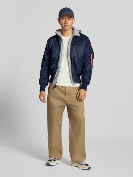 Männermodel trägt Bomberjacke mit Kapuze und Label-Details von Alpha Industries in Blau, kombiniert mit heller Hose, Ganzkörperansicht von vorne. Herren Bomberjacke Regular Fit, Reißverschluss, Eingrifftaschen, Rückenlänge 63 cm, Schulterbreite 47 cm, Brustweite 54 cm, Ärmellänge 68 cm.