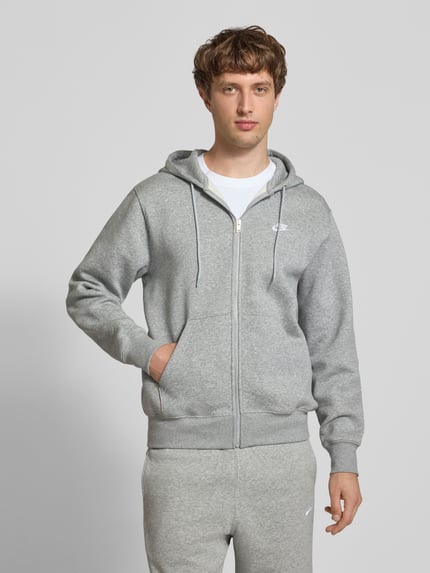 Męski model nosi Sweatjacke mit Label-Stitching und Känguru-Tasche von Nike, mieszanka bawełny, krój regular fit, kaptur, zamek błyskawiczny, kieszeń kangurka, długość tyłu w rozmiarze M 72 cm, długość rękawów w rozmiarze M 72 cm, szara, gładka tekstura, widok całej sylwetki z przodu.