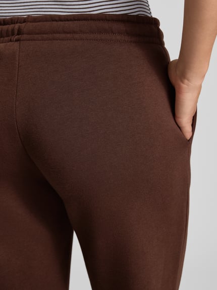 Frauenmodel trägt Wide Fit Sweatpants aus Baumwoll-Mix Modell 'Iconic' von Gina Tricot in Braun, strukturierte Oberfläche, Nahaufnahme Hüfte, Damen Sweatpants mit elastischem Bund und Tunnelzug, Innenbeinlänge bei Größe S 80 cm.