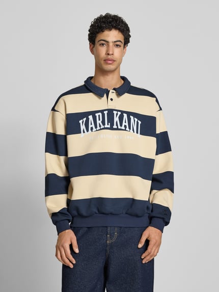 Männermodel trägt Oversized Sweatshirt mit Polokragen Modell 'Heritage' von KARL KANI im Baumwoll-Mix mit Logo-Print, navy-beige, elastische Abschlüsse, Rückenlänge bei Größe M 65 cm, Oversized-Fit, Ganzkörperansicht von vorne.