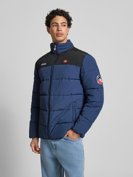 Männermodel trägt blaue Steppjacke mit schwarzen Schulterpartien, Stehkragen und Reißverschluss, Steppjacke mit Reißverschluss Modell 'NEBULA' von Ellesse, Label-Details, Eingrifftaschen, Regular Fit, Ganzkörperansicht von vorne.