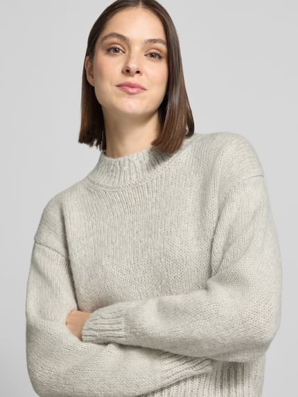 Modelka kobieca ma na sobie sweter z alpaki z prążkowanymi ściągaczami od MARC CAIN, krój regular fit, jednokolorowy, imitacja dzianiny, krótki krój, długie rękawy z prążkowanym wykończeniem, okrągły dekolt, wykończenie tułowia prążkowane, ujęcie z przodu - zbliżenie na górną część ciała.