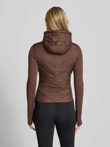 Modelka nosi kurtkę pikowaną z kapturem Model 'GRAZIA' od GUESS Activewear, krój slim fit, kolor brązowy, pełen tył, zdjęcie w widoku całej sylwetki od tyłu. Długość tyłu w rozmiarze S: 55 cm, szerokość ramion: 38 cm, szerokość w rozmiarze S: 40 cm, długość rękawów: 68 cm.
