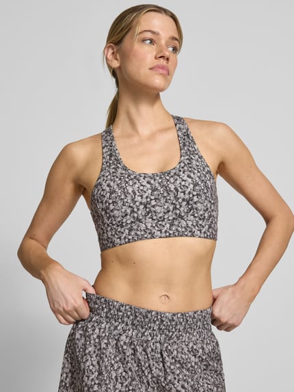 Frauenmodel trägt Freesoft Sport-BH mit Racerback Modell 'selma' von VARLEY im Material-Mix mit Stretch-Anteil und Rundhalsausschnitt, mit Logo-Print, Nahaufnahme Oberkörper.