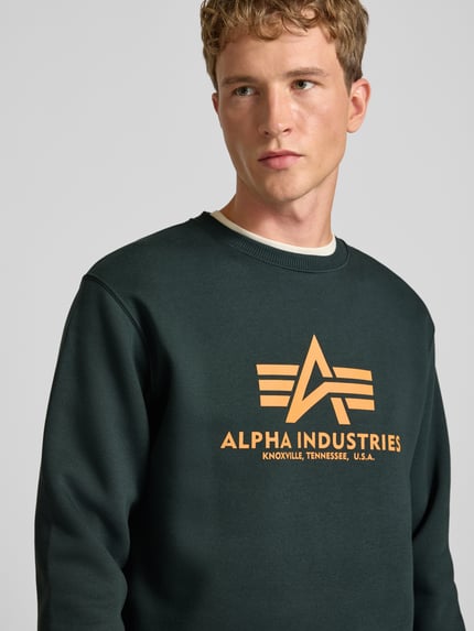 Męski model nosi Regular Fit Sweatshirt aus Baumwoll-Mix mit Logo-Print od Alpha Industries w kolorze ciemnozielonym z nadrukiem z logo, mieszanka bawełny, krój regular fit, prążkowane wykończenia, długi rękaw, okrągły dekolt, długość tyłu w rozmiarze M 67 cm, długość rękawów w rozmiarze M 64 cm, zbliżenie na górną część ciała.