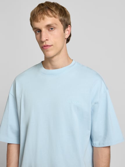 Mężczyzna model nosi Oversized T-Shirt mit Logo od REVIEW, jasnoniebieski, czysta bawełna, szeroki krój, logo, dekolt okrągły, Nahaufnahme górnej części ciała.