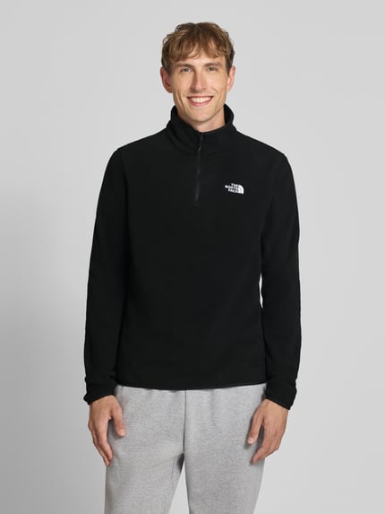 Mężczyznamodel nosi czarny sweter z polaru z kołnierzem zapinanym The North Face, Model 'GLACIER', prosty krój, wyhaftowane logo, Długość tyłu w rozmiarze M: 70 cm, Długość rękawów w rozmiarze M: 73 cm, ujęcie całej sylwetki od przodu.