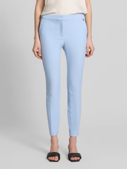 Frauenmodel trägt Slim Fit Hose mit Bügelfalten Modell 'HAGONIA-1' von HUGO in Hellblau, mit Logo-Applikation und Eingrifftaschen, Ganzkörperansicht von vorne.