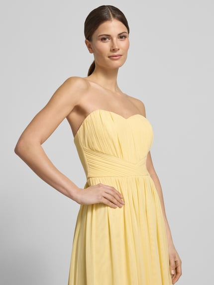 Frauenmodel trägt Cocktailkleid im Off-Shoulder-Look von Lipsy in Gelb, tailliert, fließendes Material mit Raffungen, Nahaufnahme Oberkörper.