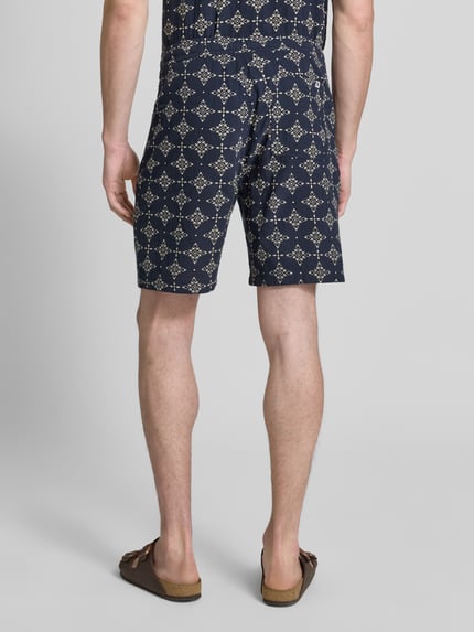 Mężczyzna nosi Relaxed Fit Stoffshorts aus einer Baumwolle od s.Oliver RED LABEL, granatowy materiał z jasnym wzorem, widok całej sylwetki z tyłu.