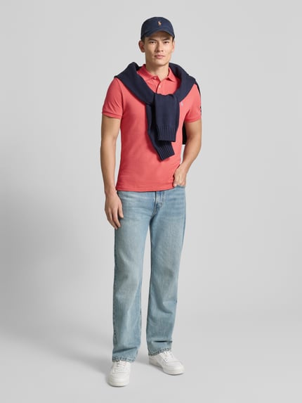 Männermodel trägt Poloshirt mit Label-Stitching von Polo Ralph Lauren in Koralle, Slim Fit, Baumwolle, kombiniert mit Jeans, Ganzkörperansicht von vorne.