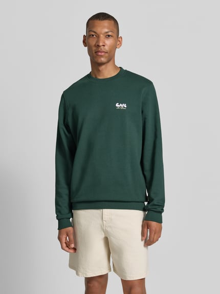 Männermodel trägt Sweatshirt mit Label-Details und Rundhalsausschnitt von Les Deux, dunkelgrün, Baumwoll-Elasthan-Mix, Regular-Fit, Ganzkörperansicht von vorne, Rückenlänge 66 cm, Ärmellänge 70 cm, Größe M.
