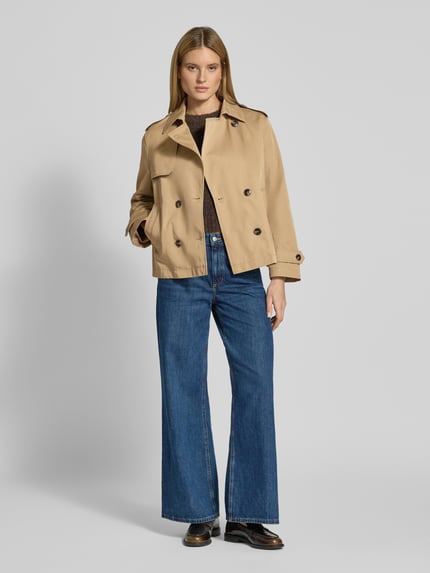 Modelka prezentuje Trenchcoat in kurzem Design mit Baumwoll-Anteil von Jake*s Casual, beżowy, prosty krój, widok całej sylwetki z przodu. Materiał mieszany z bawełną, kołnierz wykładany, dwurzędowe zapięcie, kieszenie wpuszczane, pagony na ramionach i rękawach, krótka forma. Długość pleców w rozmiarze 36: 64 cm, szerokość ramion w rozmiarze 36: 44 cm, szerokość klatki piersiowej w rozmiarze 36: 51 cm, długość rękawa w rozmiarze 36: 60 cm.