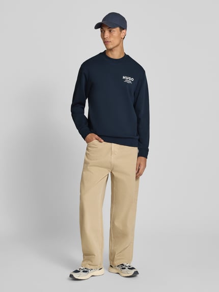 Männermodel trägt Regular Fit Sweatshirt aus reiner Baumwolle Modell 'Nespon' von HUGO Blue in Dunkelblau, beige Hose, Ganzkörperansicht von vorne.