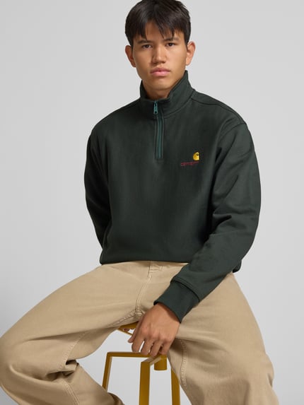 Männermodel trägt Relaxed Fit Sweatshirt aus elastischem Material-Mix von Carhartt Work In Progress in Dunkelgrün, Nahaufnahme Oberkörper. Elastischer Abschluss, Troyer-Kragen, dezentes Label-Detail, Teil des Herren-Sweatshirtsortiments.