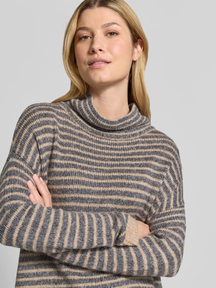 Frauenmodel trägt Oversized Fit Strickpullover mit Viskose-Anteil von Apricot, geringeltes Muster in Grau und Beige, Nahaufnahme Oberkörper. Rückenlänge 76 cm, Ärmel 41 cm, gerippte Abschlüsse, hochgeschlossener Kragen, überschnittene Schultern, verlängerte Rückseite.