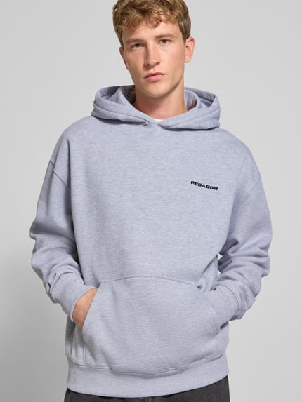 Männermodel trägt Oversized Hoodie aus Baumwoll-Mix von PEGADOR in Hellgrau mit Logo-Stickerei, Kängurutasche, Ganzkörperansicht von vorne.