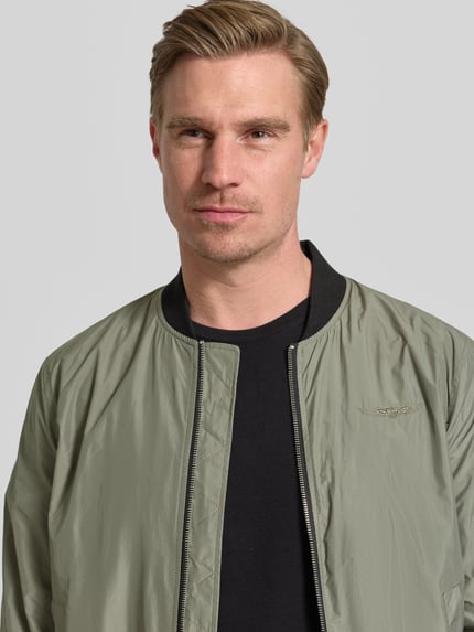 Mężczyzna model nosi Bomberjacke mit Label-Patch und Eingrifftaschen od Original Bombers, jasny oliwkowy kolor, tekstura gładka, zamek błyskawiczny, detal naszywki, szerokość w rozmiarze M: 53 cm, krój regular fit, zbliżenie na górną część ciała