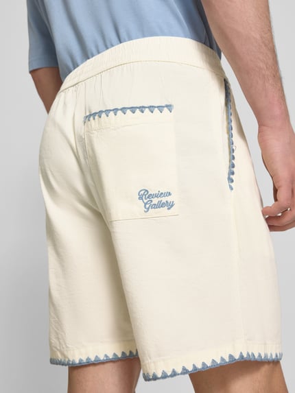 Männermodel trägt Shorts mit umstochener Kante von REVIEW, Baumwoll-Mix in Creme mit blauen Details, Bund und Tasche, Ganzkörperansicht Rückansicht.