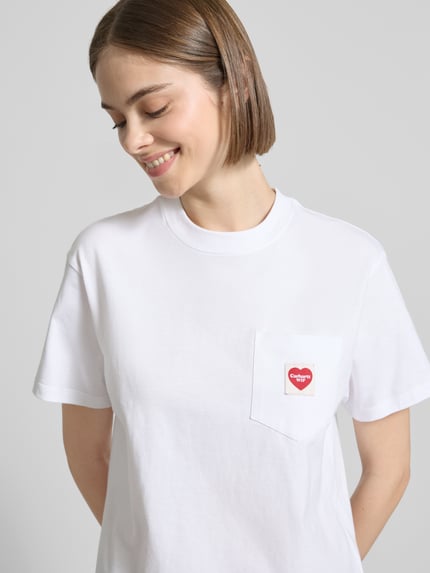 Frauenmodel trägt Damen T-Shirt mit Label-Patch von Carhartt Work In Progress in Weiß, Reine Baumwolle, Gerader-Schnitt, Rundhalsausschnitt, Brusttasche, Rückenlänge bei Größe S 58 cm, Nahaufnahme Oberkörper.