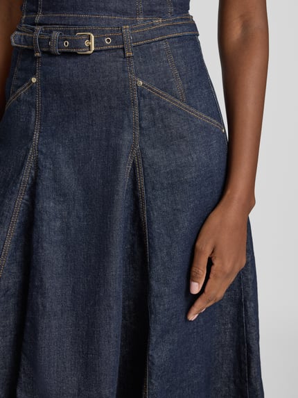 Kobieta nosi spódnicę jeansową z paskiem Model 'RAYNE' od GUESS, ciemnoniebieski denim, wpuszczane kieszenie, Mieszanka bawełny z dodatkiem streczu, Linia a, Spódnica midi, sprzączka, szwy w kontrastowym kolorze, zapięcie na haftkę i zamek błyskawiczny, Szerokość w rozmiarze 27: 37 cm, całkowita długość w rozmiarze 27: 82 cm, zbliżenie na górną część spódnicy.
